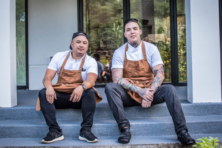 Chef Collantes and Chef Cha sit on steps outdoors in their aprons.