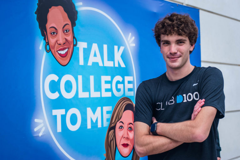 Dykan Carollo of D100 Radio poses in front of a Talk College To Me Podcast poster.