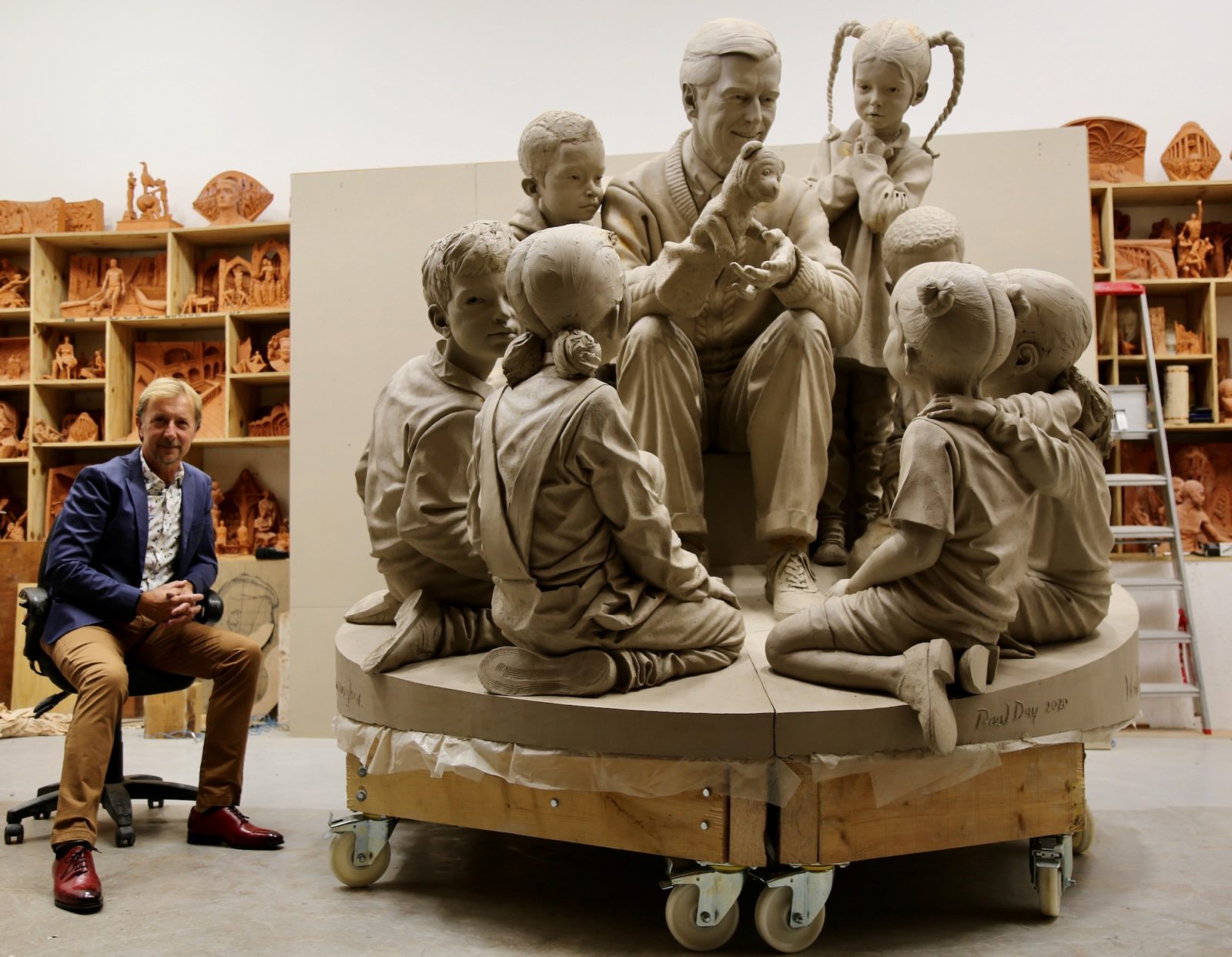 Paul day sits next to "A Beautiful for a Neighbor" statue featureing Mister Rogers, his puppets, and children.
