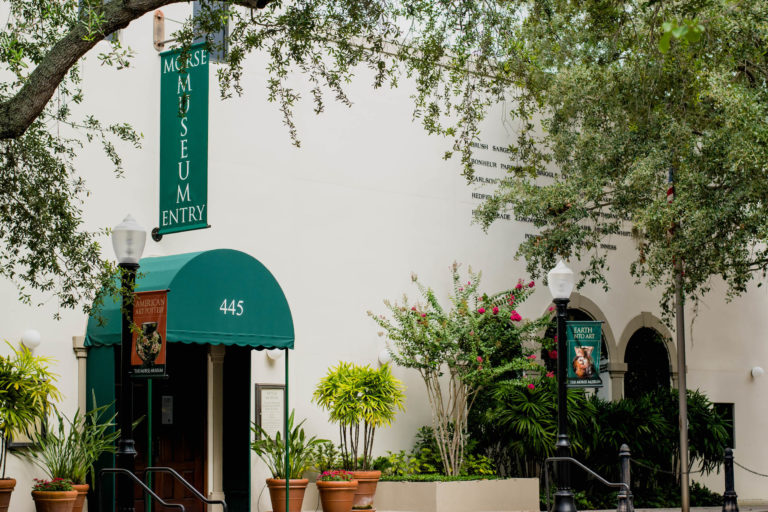 Photo shows white building with dark green sign and covering with green trees