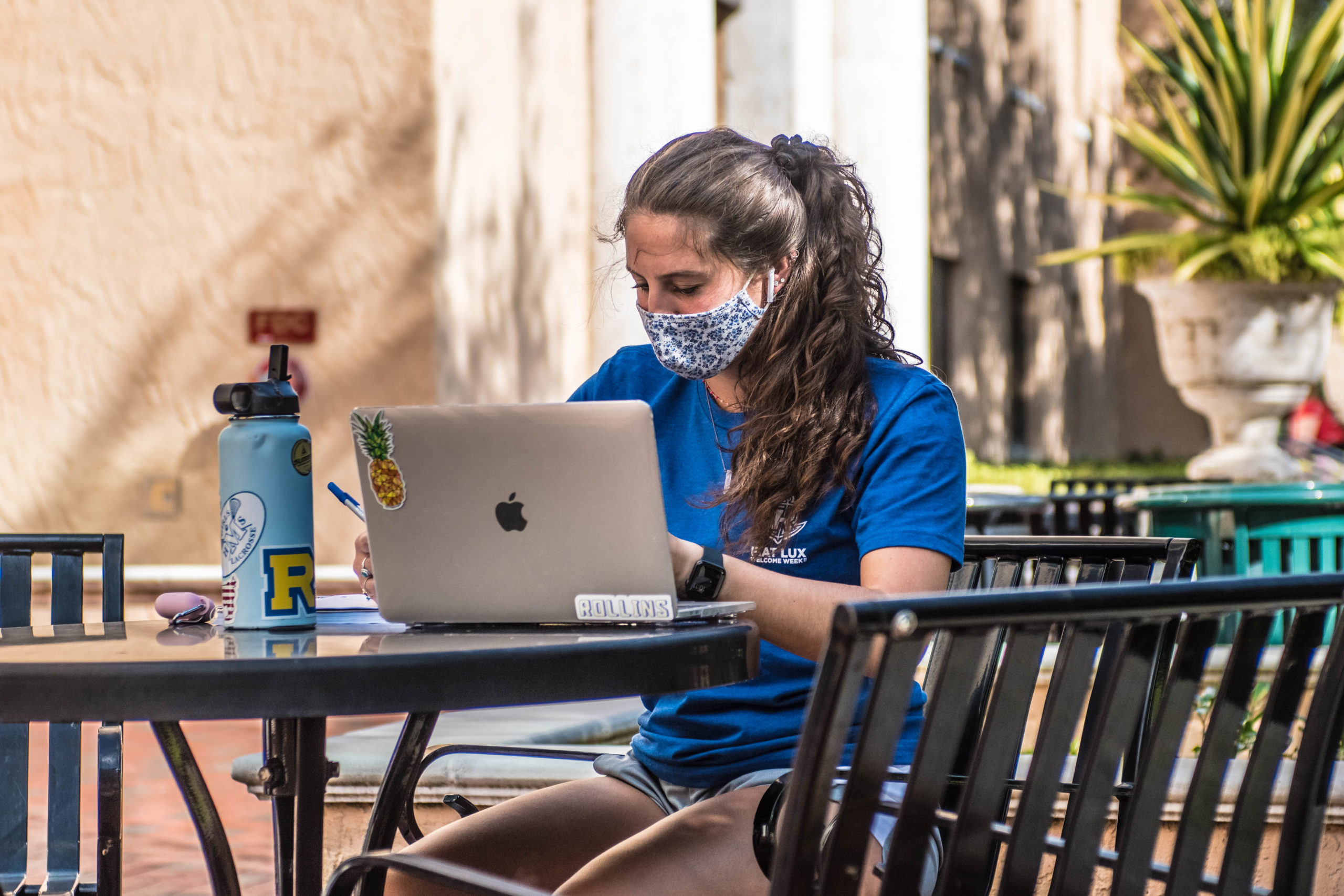 Rollins student sits masked outdoors on her laptop.