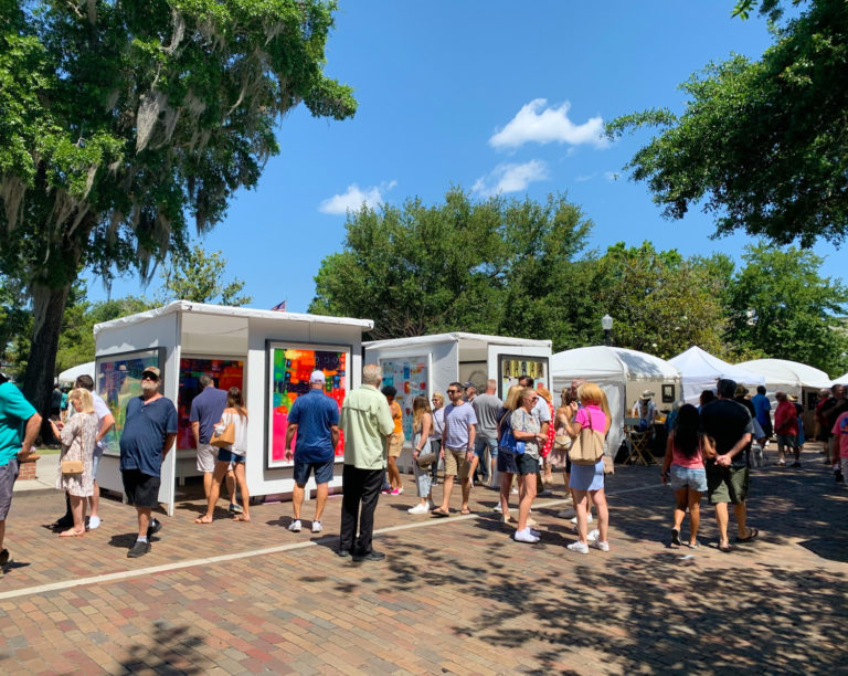 Image shows white tents on Park avenue, filled with colorful art and people shopping.