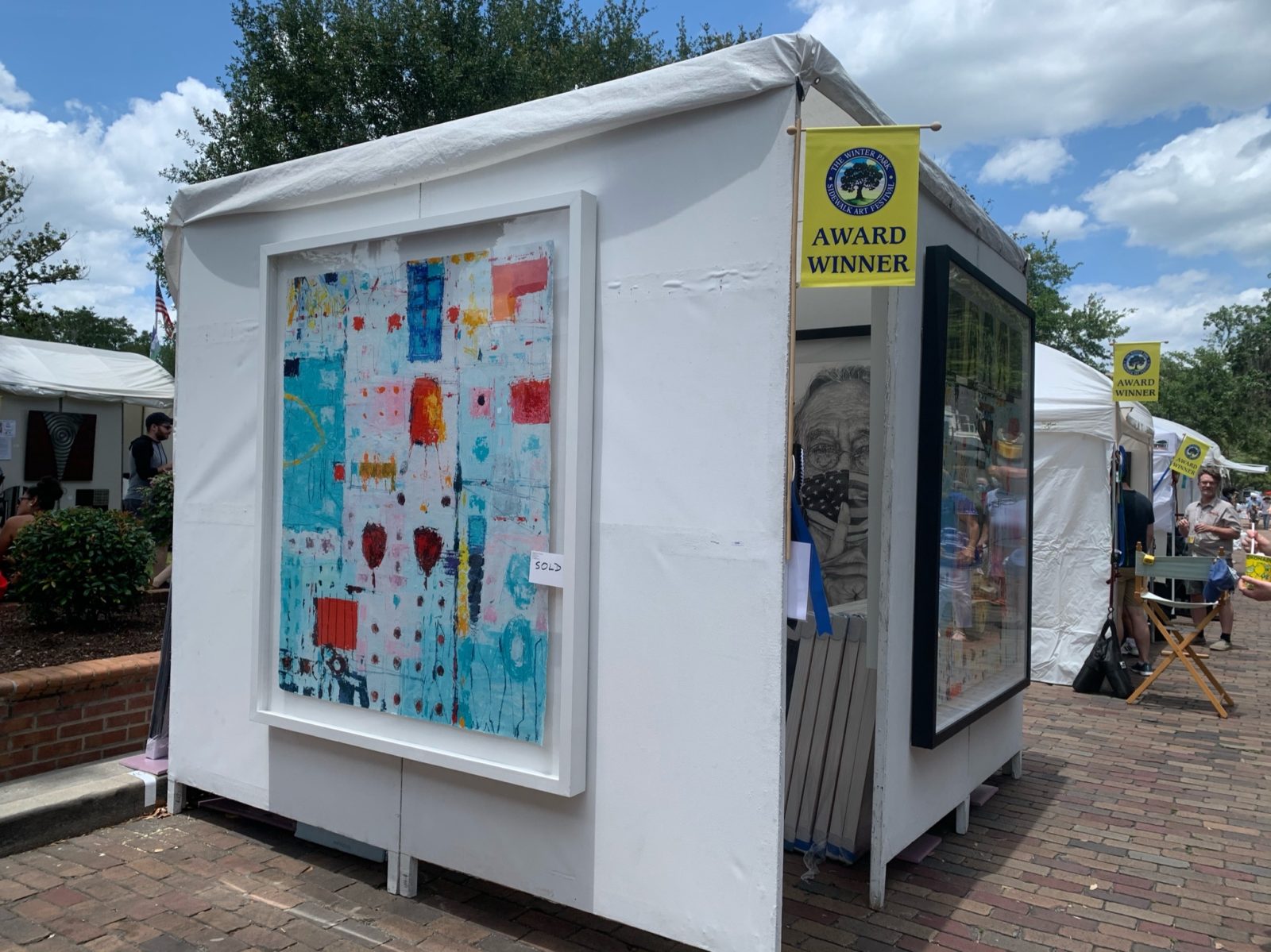 White booth from the Sidewalk Art Festival shows a painting with blue, red, orange, yellow and dark blue colors organized into an abstract painting.