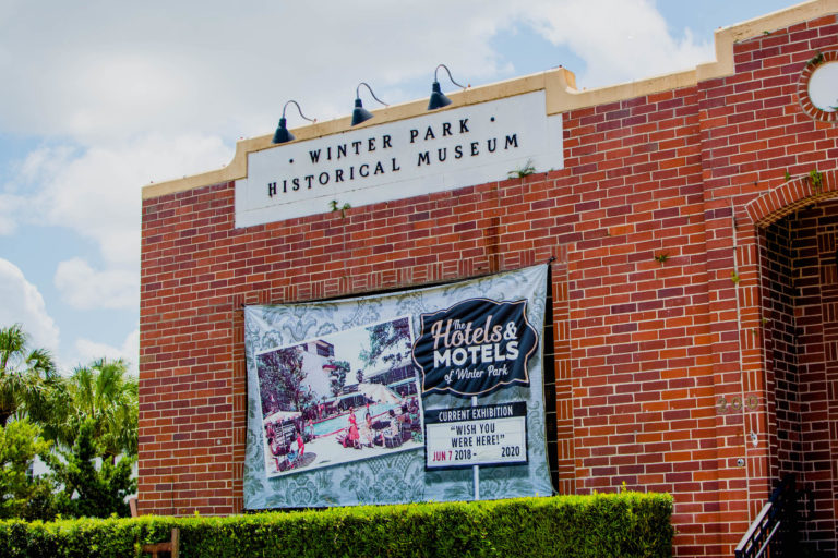 Photo shows red brick building with green hedge and white sign in front of a blue sky