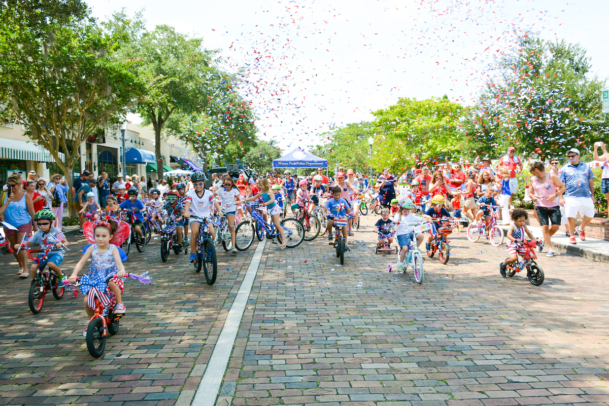 Many people riding bikes on a grey paved road celebrate the Fourth of July with red, white, and blue confetti.