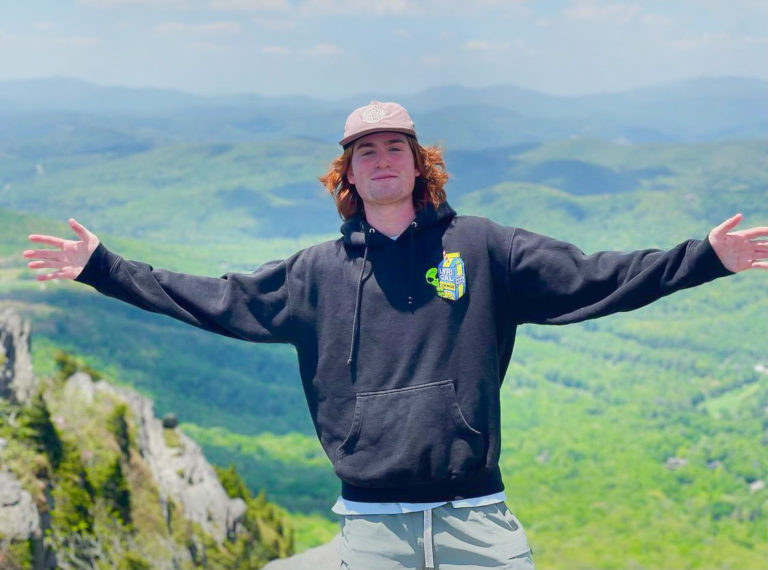 A tall boy with red hair stands on a mountain next to a green park.