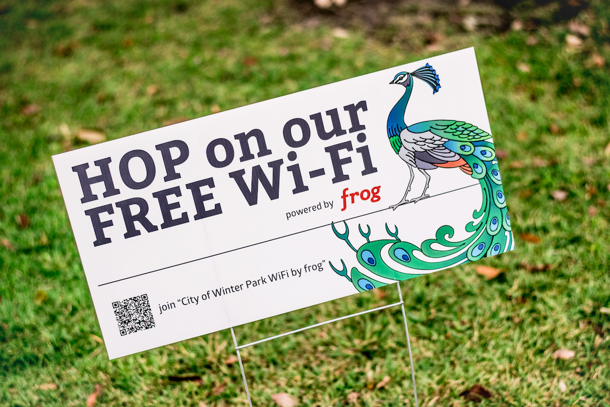 A white sign that says "Hop on our free Wi-Fi" has a blue peacock on it in green grass